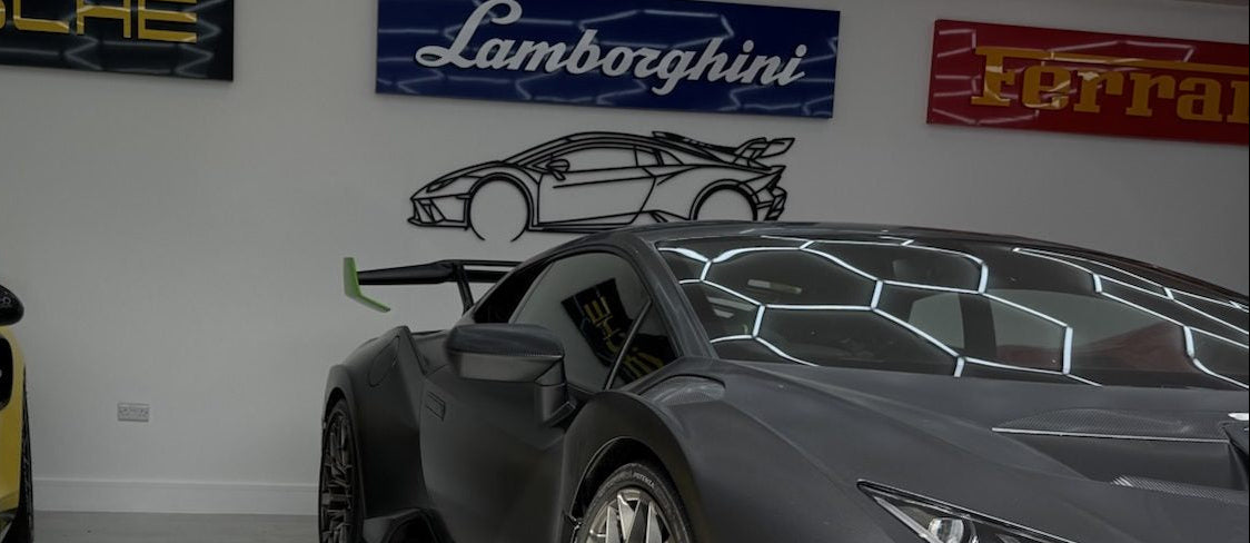 Two sports cars, one yellow and one gray, in a showroom with Porsche, Lamborghini, and Ferrari logos on the wall.